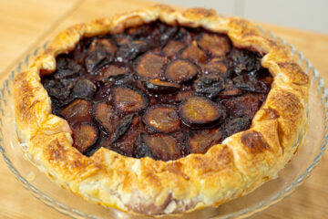 Uma tarte rústica e dourada sobre um prato, com um rebordo de massa folhada crocante e polvilhado com açúcar que se dobra irregularmente sobre um centro vibrante de metades de ameixa vermelha assadas e brilhantes.