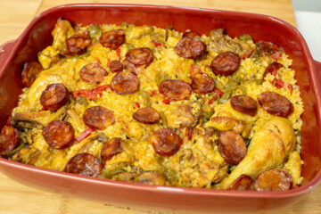 Travessa rústica de barro cheia de arroz carolino cozinhado no forno, com uma tonalidade alaranjada do açafrão, coberto com pedaços de frango do campo dourados, pimentos e rodelas de chouriço tostadas.