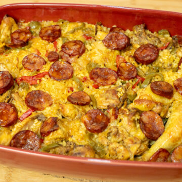 Travessa rústica de barro cheia de arroz carolino cozinhado no forno, com uma tonalidade alaranjada do açafrão, coberto com pedaços de frango do campo dourados, pimentos e rodelas de chouriço tostadas.