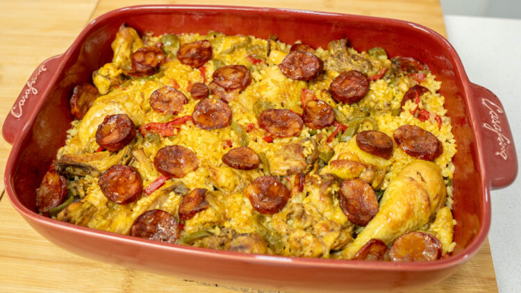 Travessa rústica de barro cheia de arroz carolino cozinhado no forno, com uma tonalidade alaranjada do açafrão, coberto com pedaços de frango do campo dourados, pimentos e rodelas de chouriço tostadas.