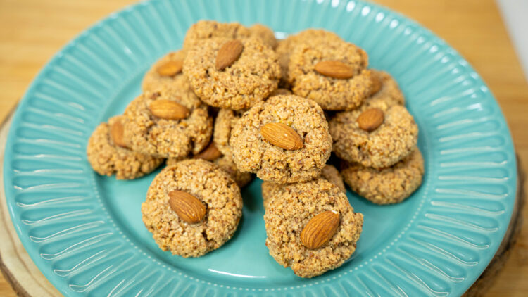 Tabuleiro com biscoitos de amêndoa rústicos e escuros, acabados de cozer no forno, decorados com uma amêndoa inteira no centro de cada um.