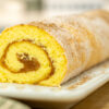Torta enrolada de pão de ló e maçã colocada numa travessa de servir, exibindo uma massa dourada e fofa polvilhada com açúcar e canela, deixando ver o recheio cremoso de maçã no interior da espiral.