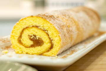 Torta enrolada de pão de ló e maçã colocada numa travessa de servir, exibindo uma massa dourada e fofa polvilhada com açúcar e canela, deixando ver o recheio cremoso de maçã no interior da espiral.