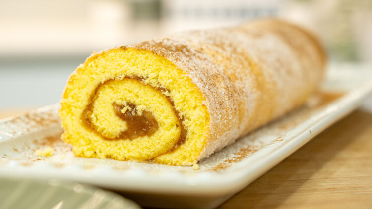 Torta enrolada de pão de ló e maçã colocada numa travessa de servir, exibindo uma massa dourada e fofa polvilhada com açúcar e canela, deixando ver o recheio cremoso de maçã no interior da espiral.