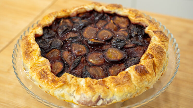 Uma tarte rústica e dourada sobre um prato, com um rebordo de massa folhada crocante e polvilhado com açúcar que se dobra irregularmente sobre um centro vibrante de metades de ameixa vermelha assadas e brilhantes.