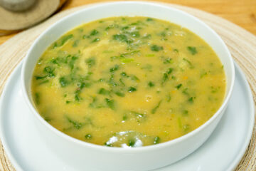 Tigela de louça branca com sopa de nabiças quente e fumegante, destacando o creme alaranjado salpicado com folhas verdes de nabiça e grãos inteiros de feijão manteiga, regada com um fio de azeite.