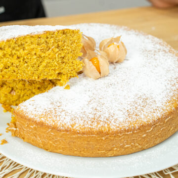 Bolo de abóbora caseiro grande e alto num prato de servir branco, generosamente polvilhado com açúcar em pó e decorado com três fisális e as suas folhas secas no topo.