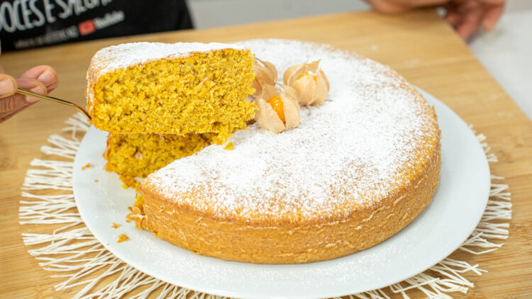 Bolo de abóbora caseiro grande e alto num prato de servir branco, generosamente polvilhado com açúcar em pó e decorado com três fisális e as suas folhas secas no topo.
