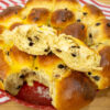 Travessa de barro redonda contendo pãezinhos doces dourados e brilhantes, unidos num grande pão brioche, pontilhados por pepitas de chocolate derretidas, com uma textura visivelmente macia