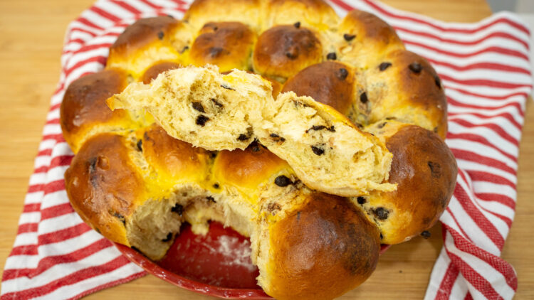 Travessa de barro redonda contendo pãezinhos doces dourados e brilhantes, unidos num grande pão brioche, pontilhados por pepitas de chocolate derretidas, com uma textura visivelmente macia