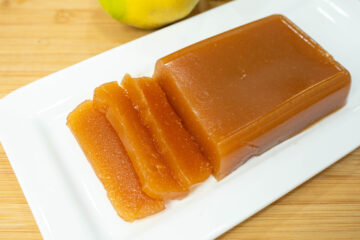 Taças cheias de marmelada caseira firme, acompanhadas por frascos de vidro invertidos contendo geleia de marmelo com uma cor âmbar vibrante e translúcida.