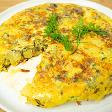 Uma tortilha dourada e apetitosa, servida num prato plano, decorada com um raminho de salsa fresca no topo, com o interior visivelmente recheado de cogumelos, presunto e queijo.