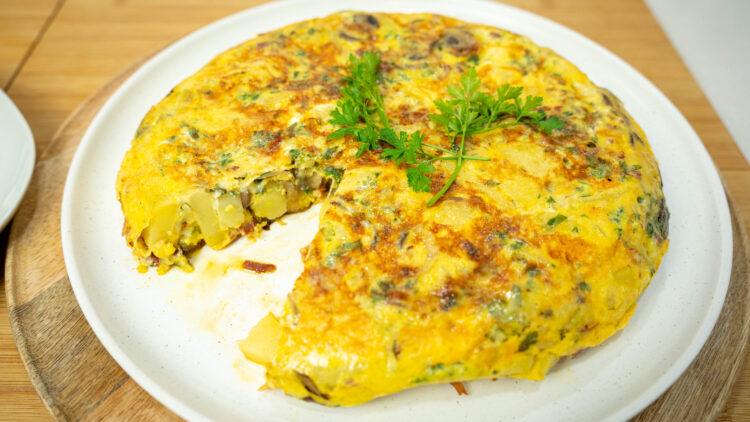 Uma tortilha dourada e apetitosa, servida num prato plano, decorada com um raminho de salsa fresca no topo, com o interior visivelmente recheado de cogumelos, presunto e queijo.