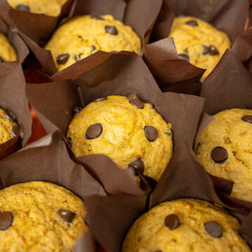 Muffins de abóbora caseiros e dourados dispostos numa travessa laranja temática, com pepitas de chocolate visíveis no topo e uma textura visivelmente fofa.